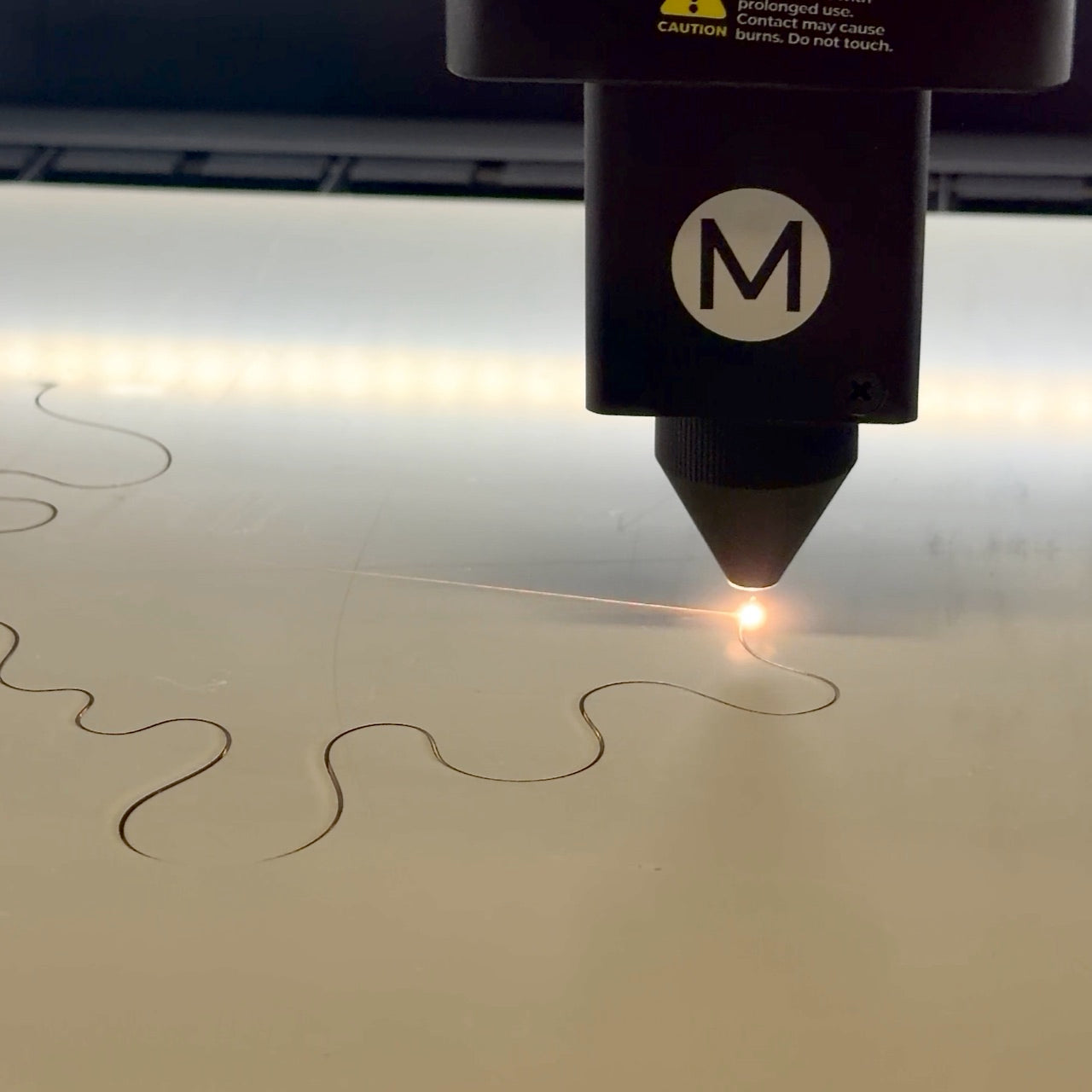 Laser engraving machine in action on a piece of wood with a visible 'M' brand logo.